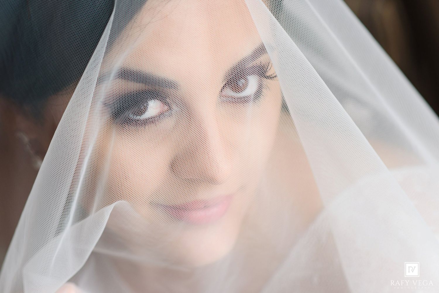 Bride with dramatic smoky eye makeup gazing through a sheer white wedding veil in a close-up portrait photo.