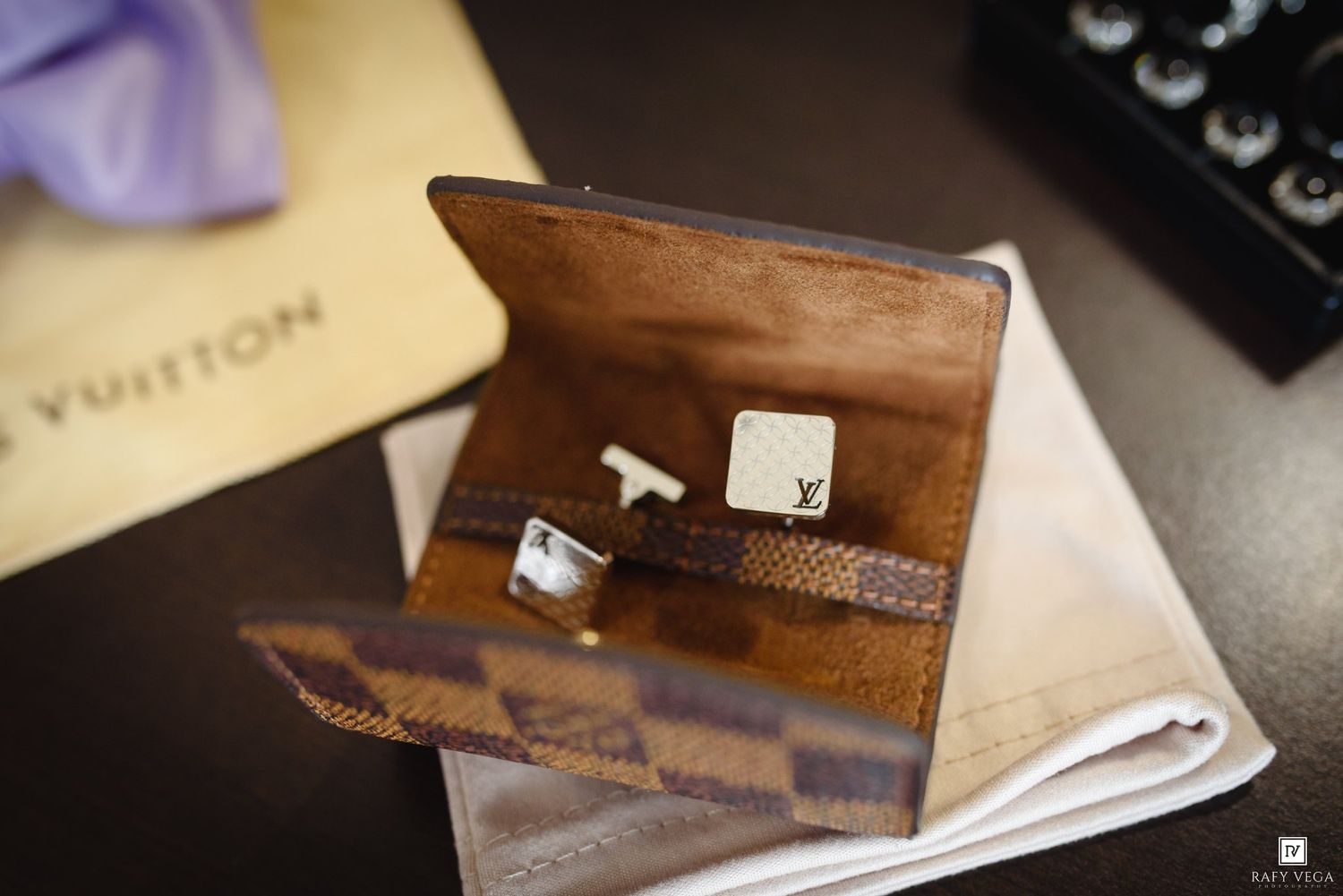 Louis Vuitton damier cufflink case open displaying silver cufflinks on white cloth with purple tie in background.
