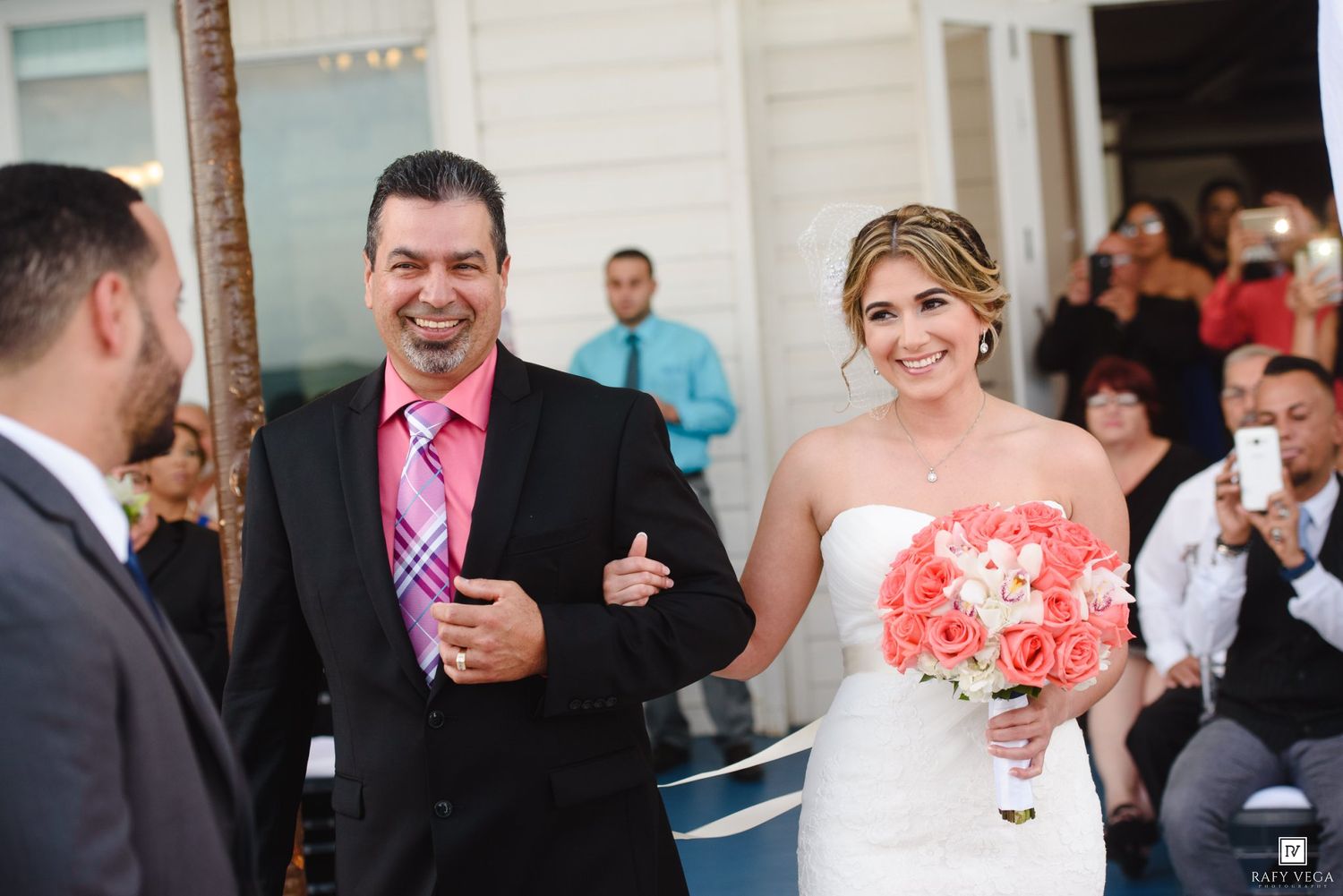 Bride holding pink bouquet walks down aisle with escort at outdoor wedding ceremony.