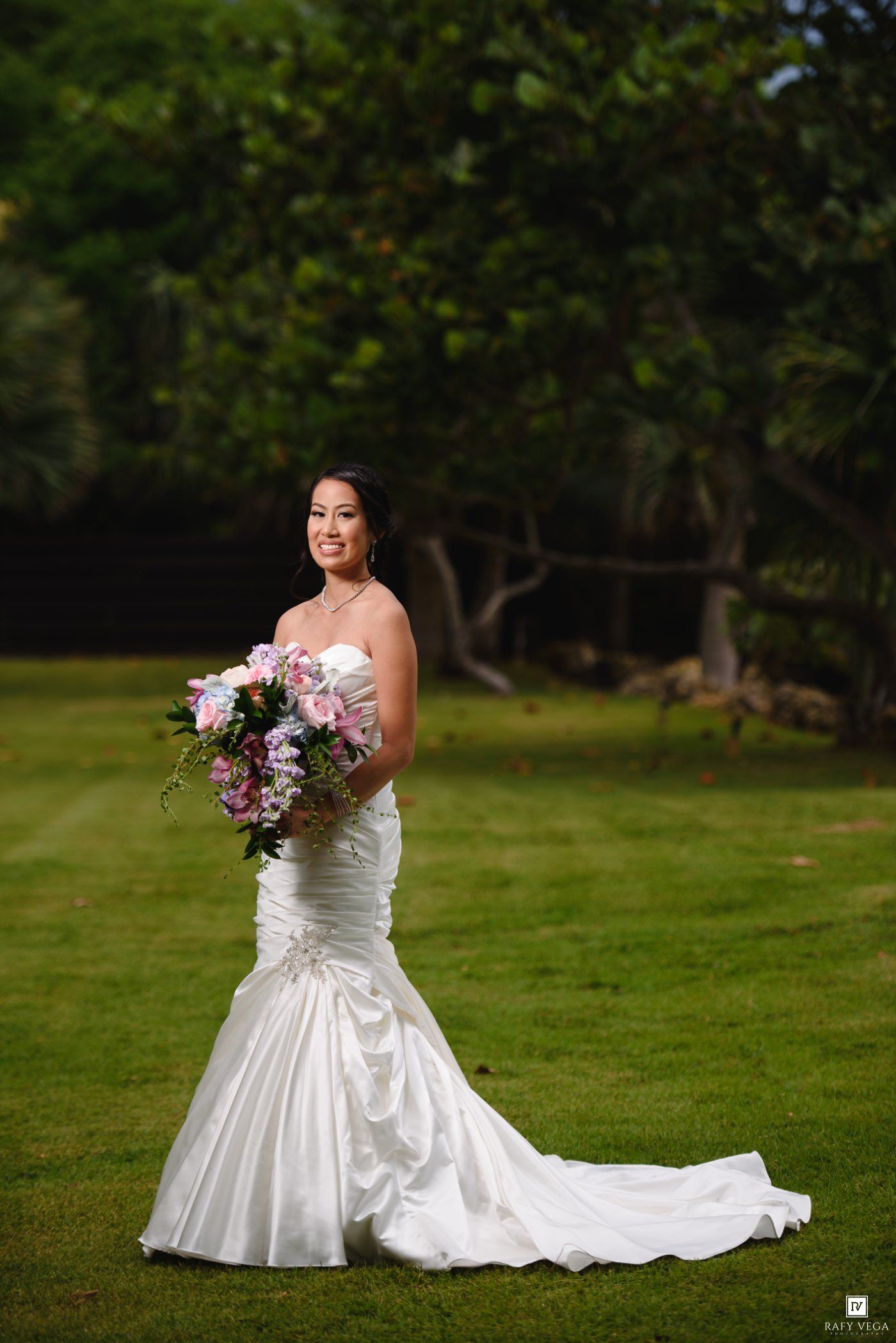 Bride in white mermaid gown holding pink floral bouquet stands on green lawn with lush trees backdrop.