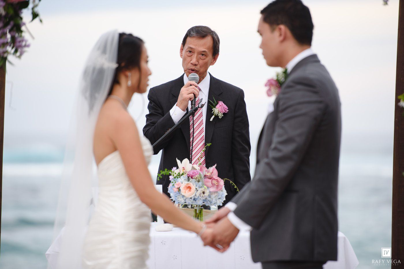 Officiant speaks at outdoor beach wedding ceremony as bride holds colorful bouquet facing groom in gray suit.