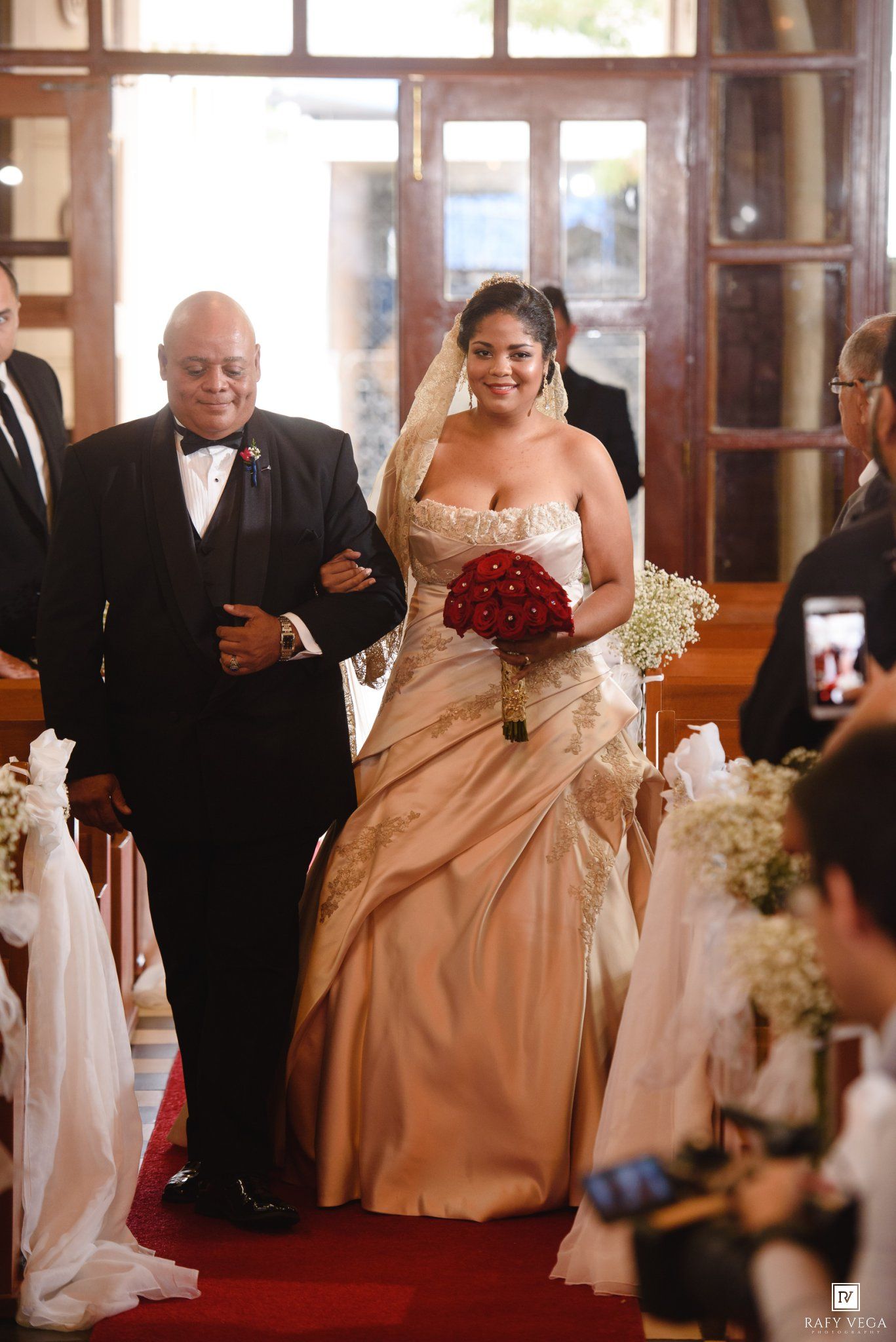 Bride in gold strapless gown walks down church aisle holding red bouquet, escorted by man in tuxedo.