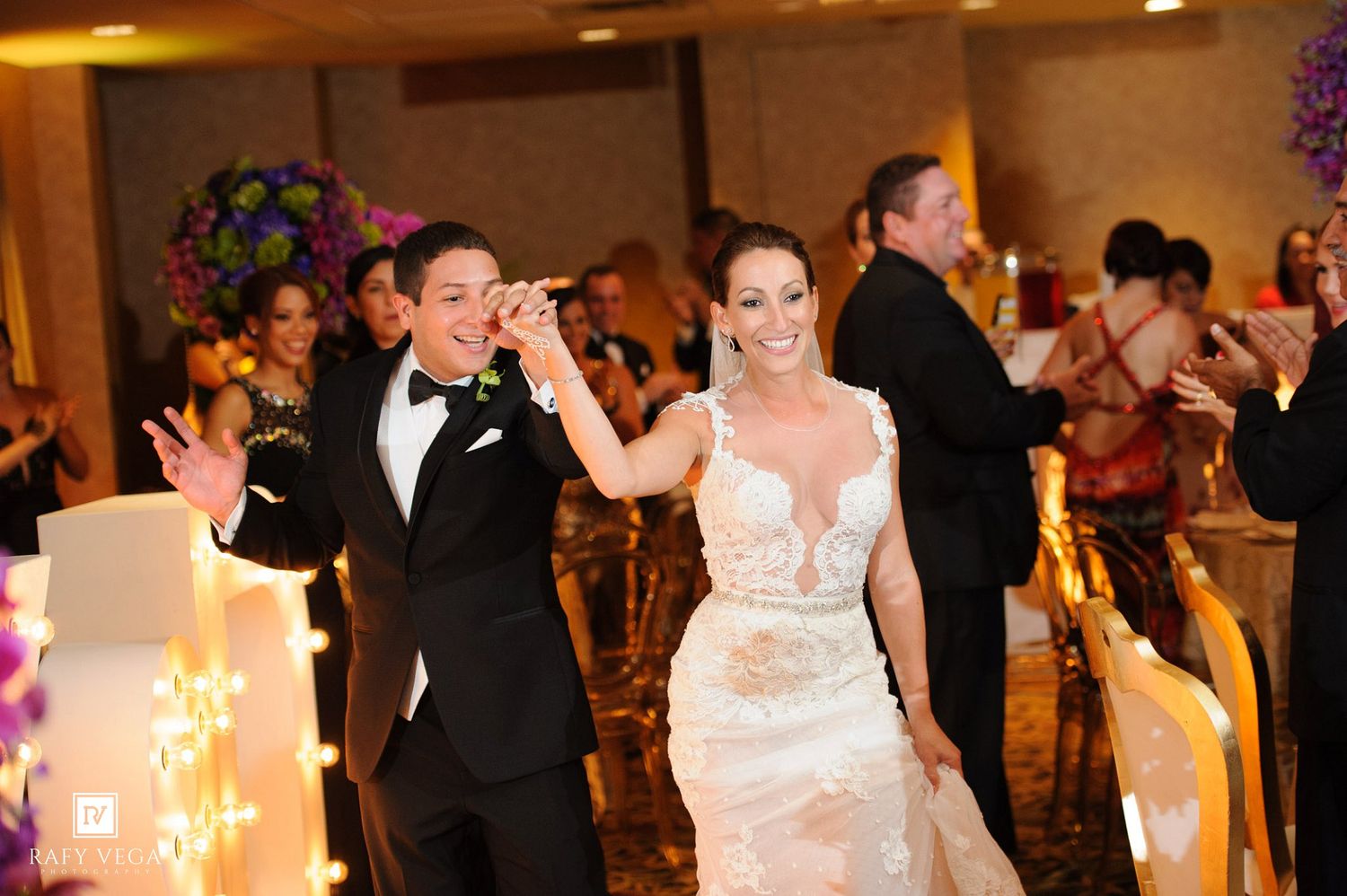 Bride and groom entering wedding reception, smiling joyfully, guests applauding in elegant ballroom.