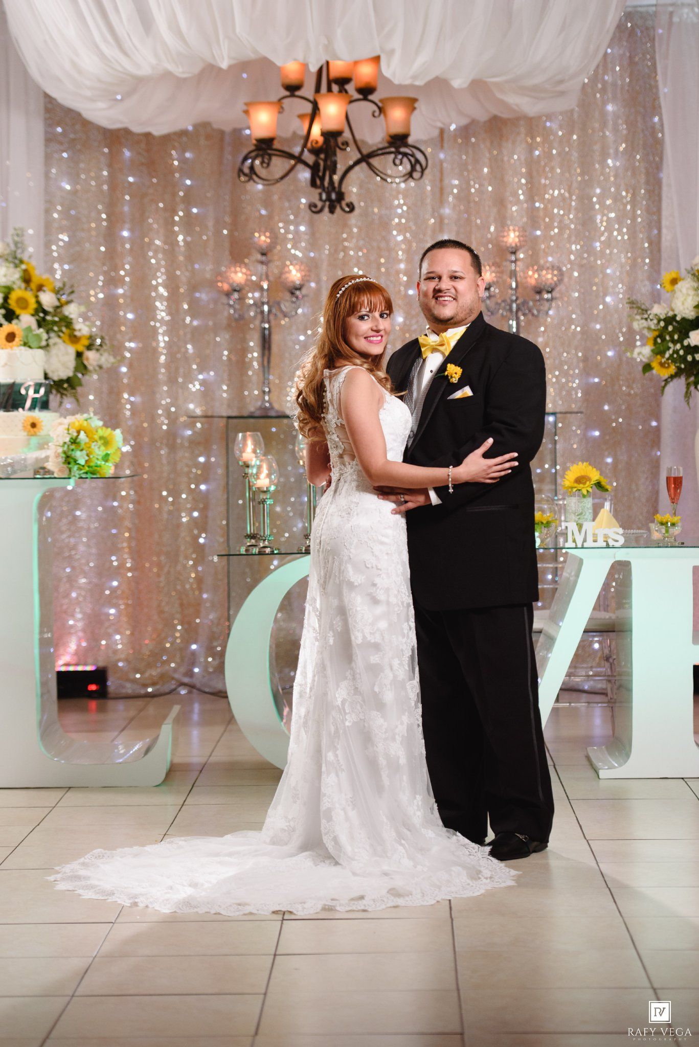 Bride in white lace gown and groom in black tuxedo with yellow boutonniere pose before sparkling backdrop at wedding reception.
