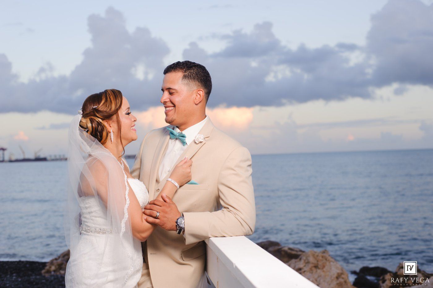 A bride and groom share a romantic moment by the ocean at sunset, with the groom in a tan suit and bow tie.