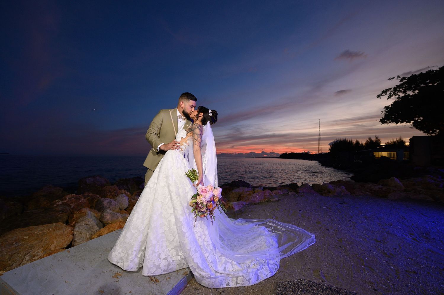 Bride and groom share a romantic kiss at sunset by the ocean, with colorful sky and rocky shoreline backdrop.