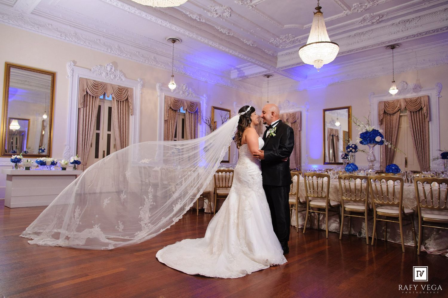 Bride and groom share a romantic kiss in an elegant ballroom, her long lace veil flowing dramatically beside them.