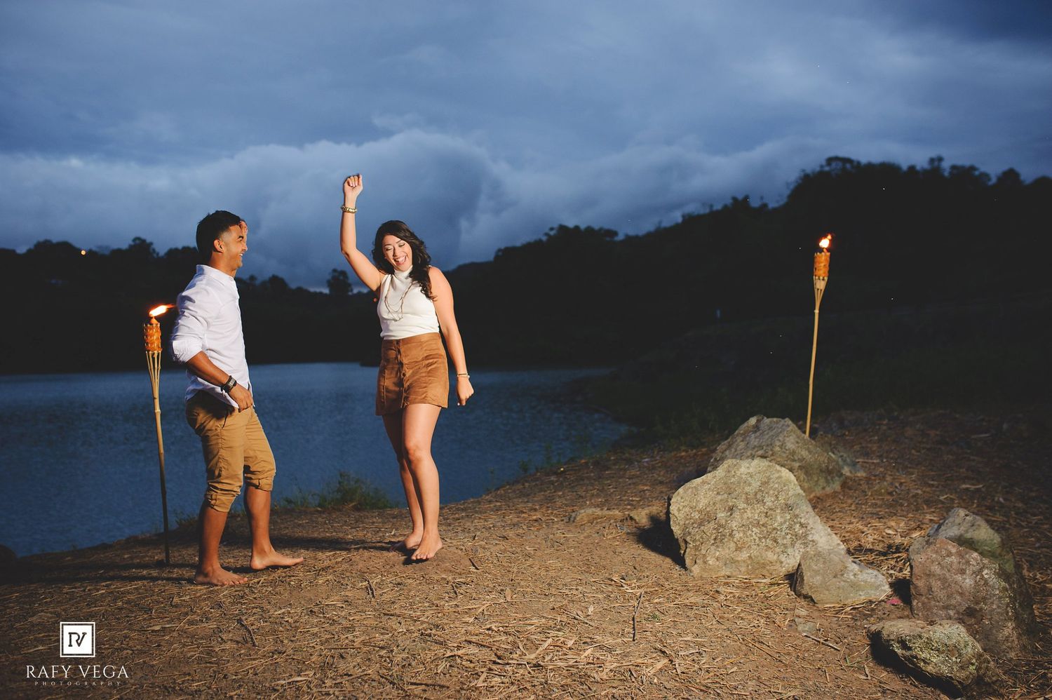 Couple dancing romantically by a lakeside at dusk with a lit torch and stormy sky in the background.