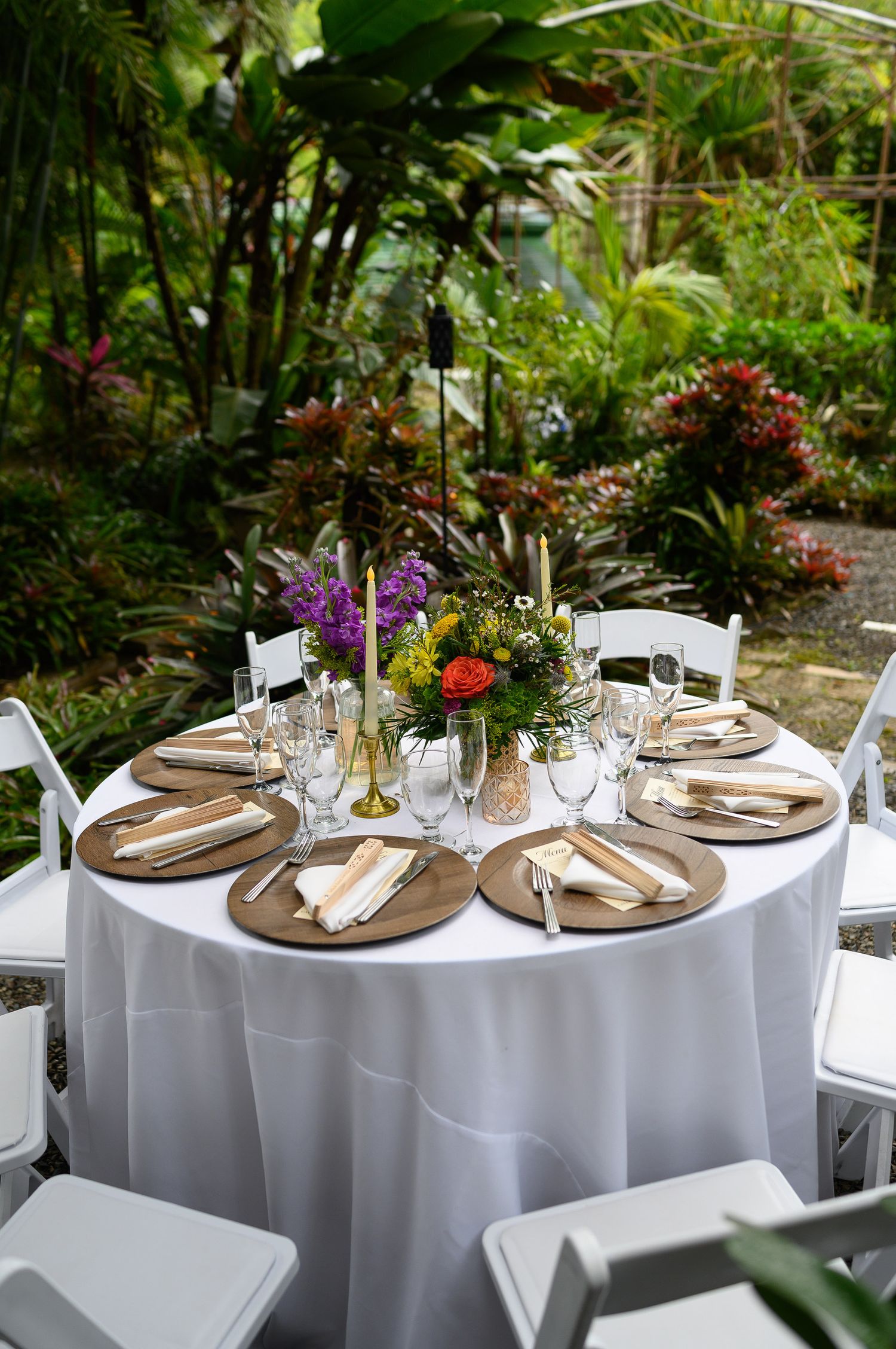 Elegant outdoor wedding reception table with white linen, wooden chargers, floral centerpiece, and lush tropical garden backdrop.