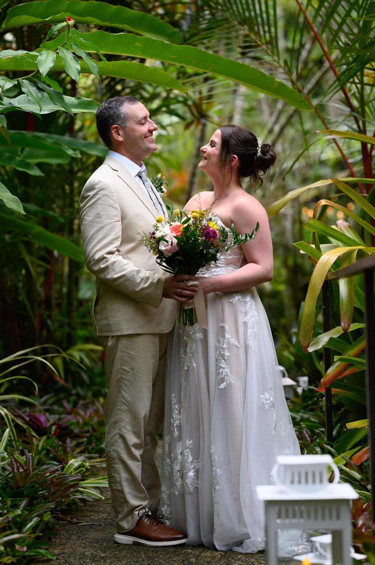 Bride and groom share a romantic moment in a lush tropical garden, bride holding a colorful bouquet in a white gown.