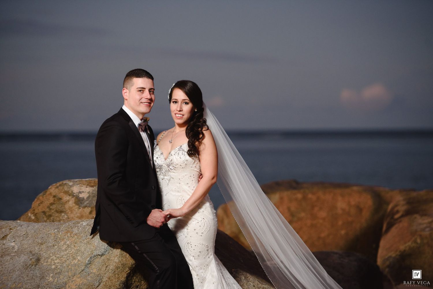 Bride and groom posing on rocky shoreline at dusk, bride in lace gown with veil, groom in black tuxedo.