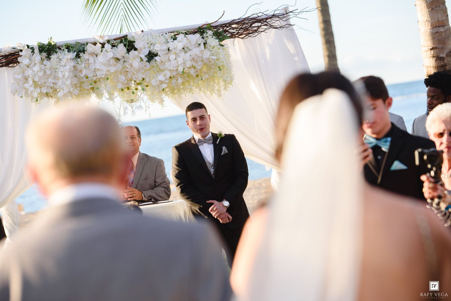 Groom stands at beach wedding altar adorned with white flowers, awaiting bride during outdoor ceremony.
