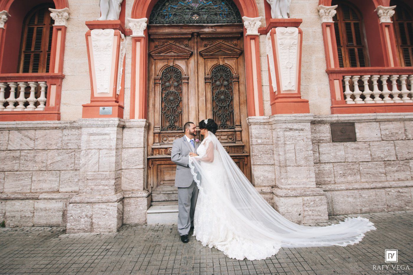 Bride and groom pose in front of an ornate red building with grand wooden doors on their wedding day.