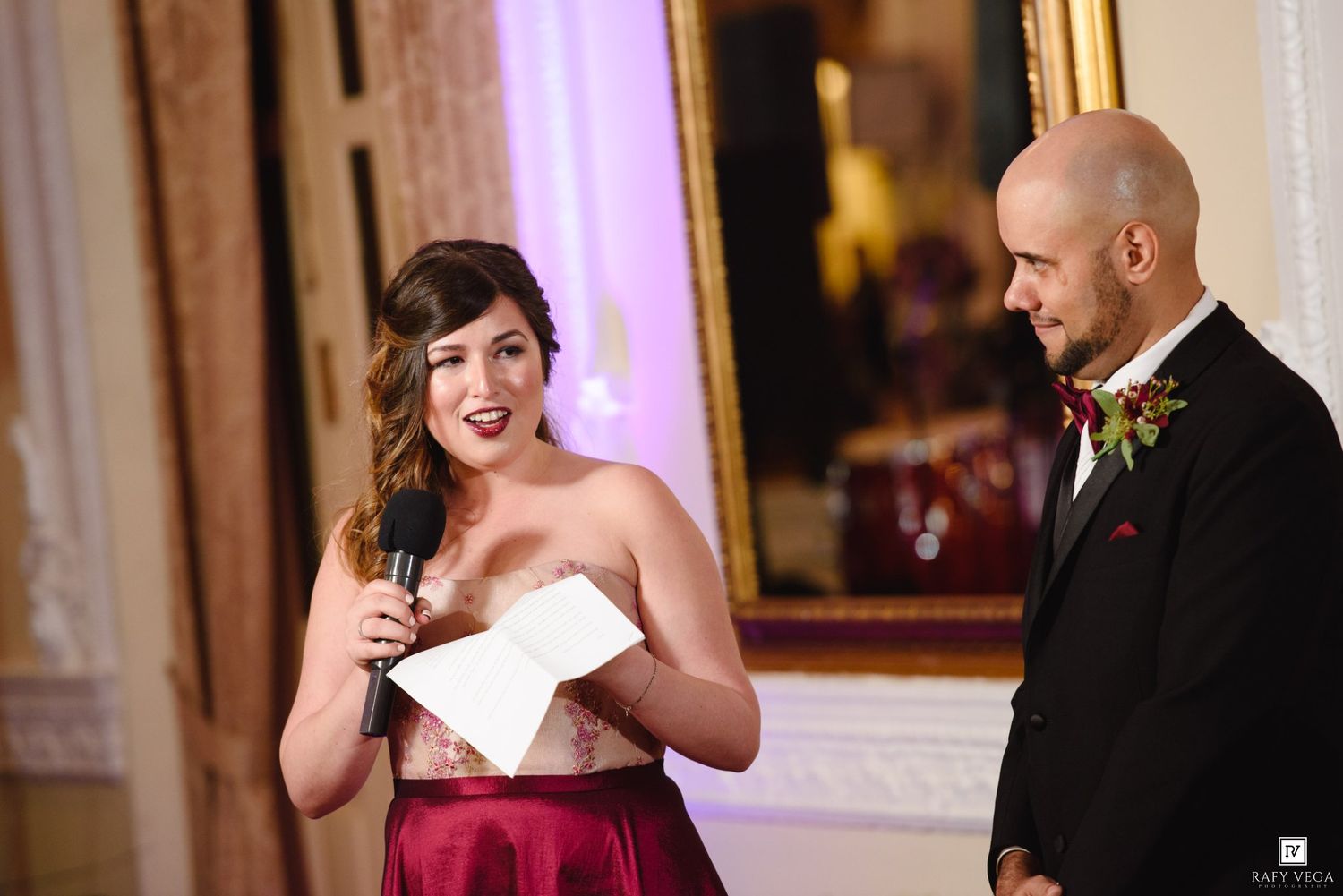 Woman in red gown speaks at wedding reception while bald man in tuxedo listens in elegant ballroom.