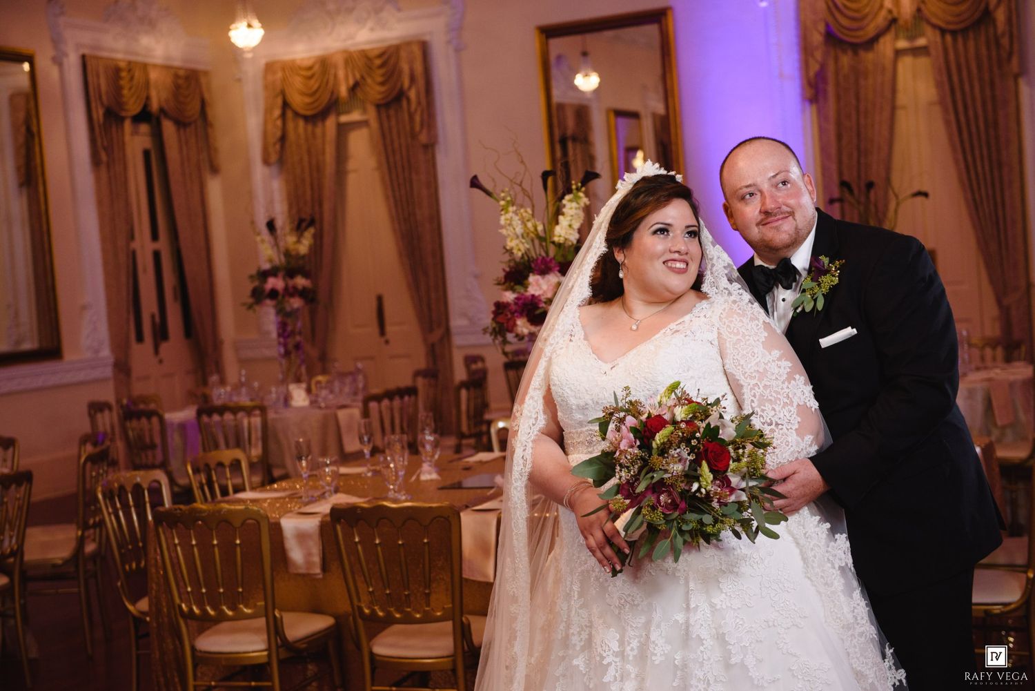 Bride in lace gown holding bouquet stands with groom in tuxedo in elegant gold-accented ballroom wedding reception.