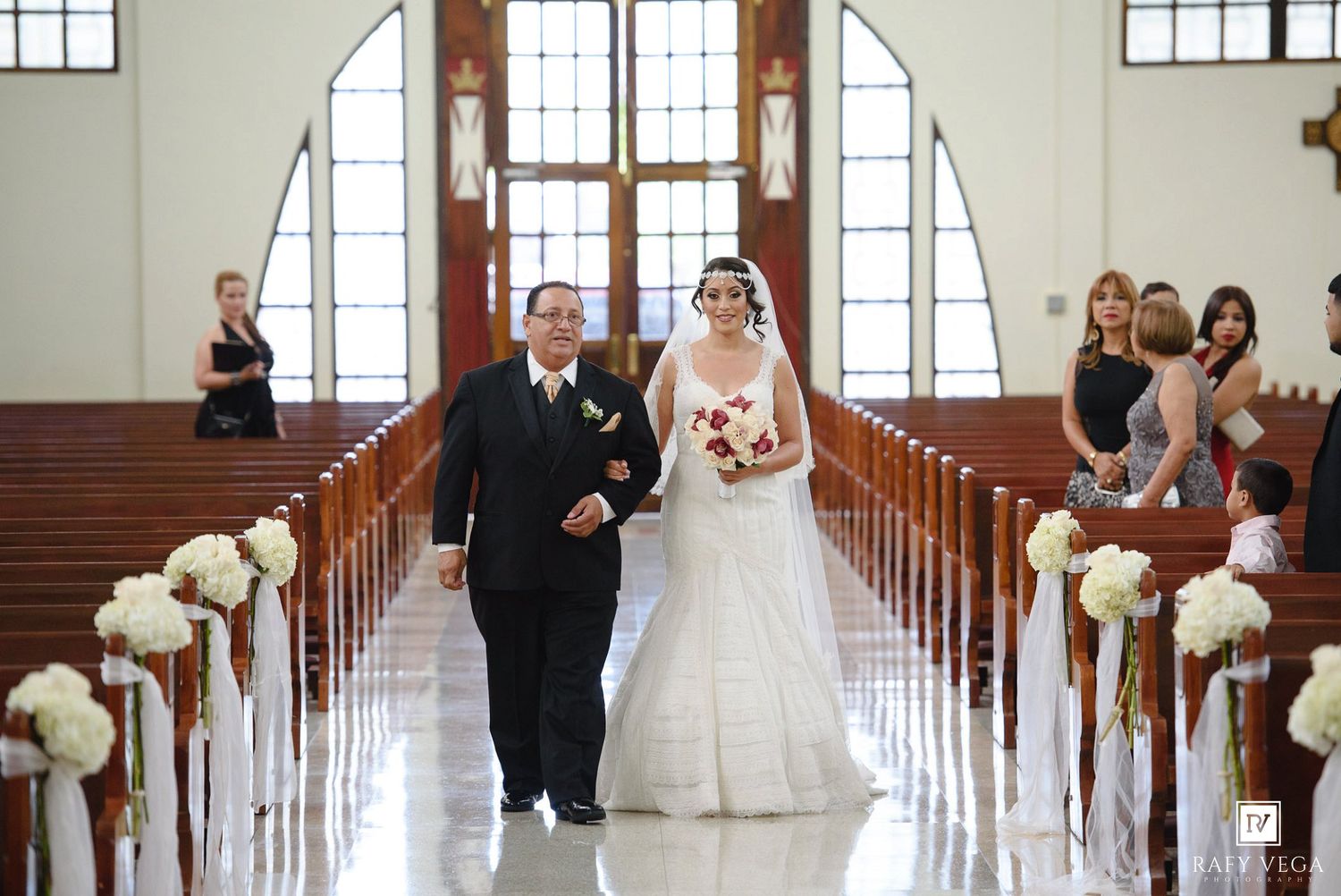 Bride walks down church aisle with escort, wearing white gown, holding red bouquet, pews decorated with white floral arrangements.