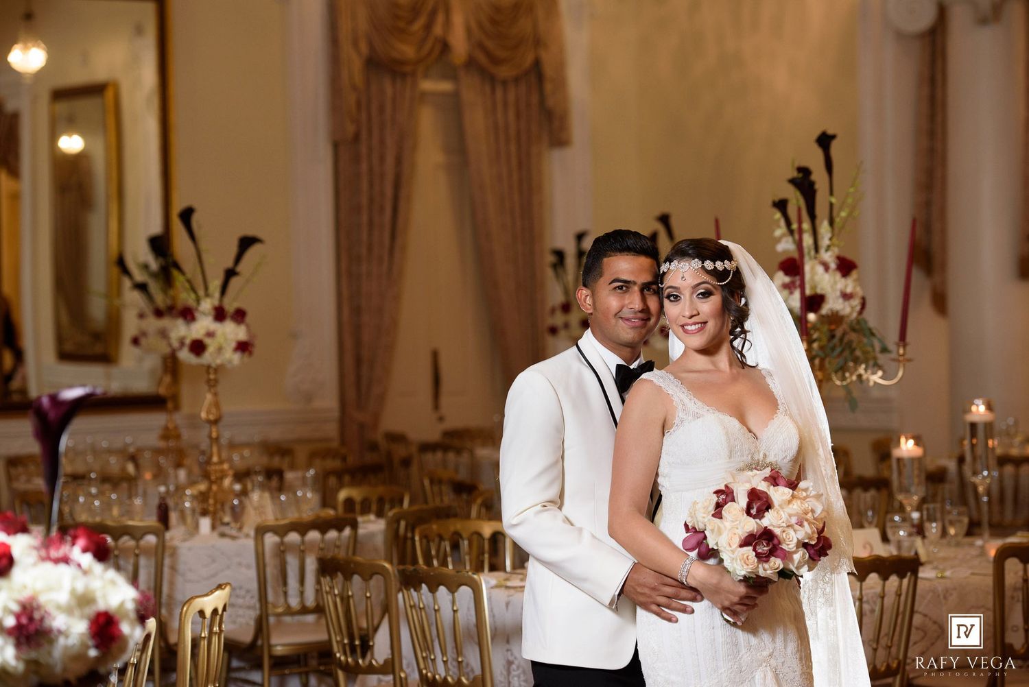 Elegant bride and groom posing in a grand ballroom with gold chairs and floral arrangements on their wedding day.