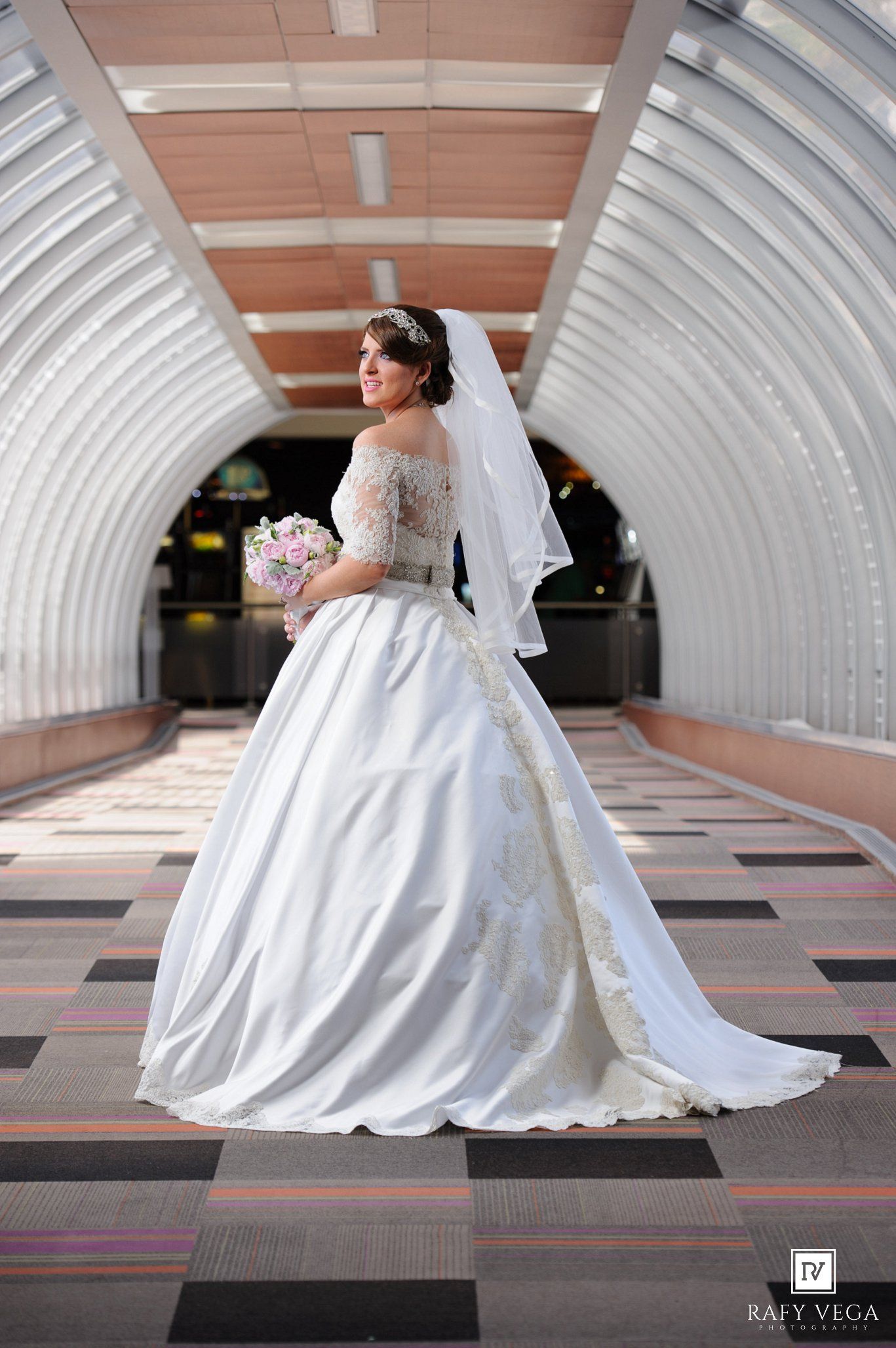 Bride in elegant white ball gown with lace sleeves and veil holds bouquet in arched tunnel corridor.