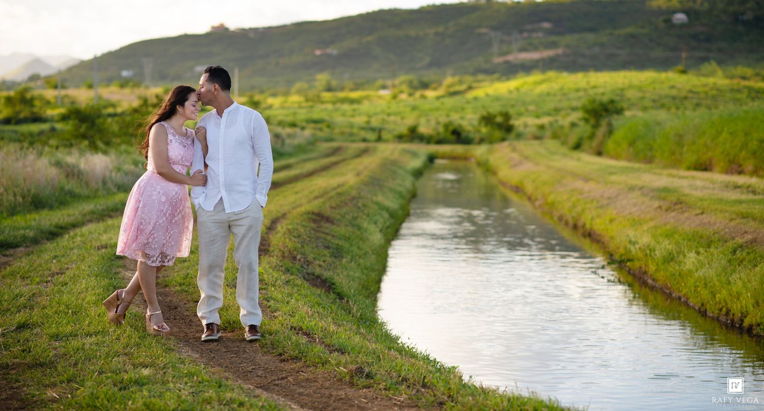 Couple sharing a romantic moment on a scenic green pathway beside a serene canal with lush hills backdrop.