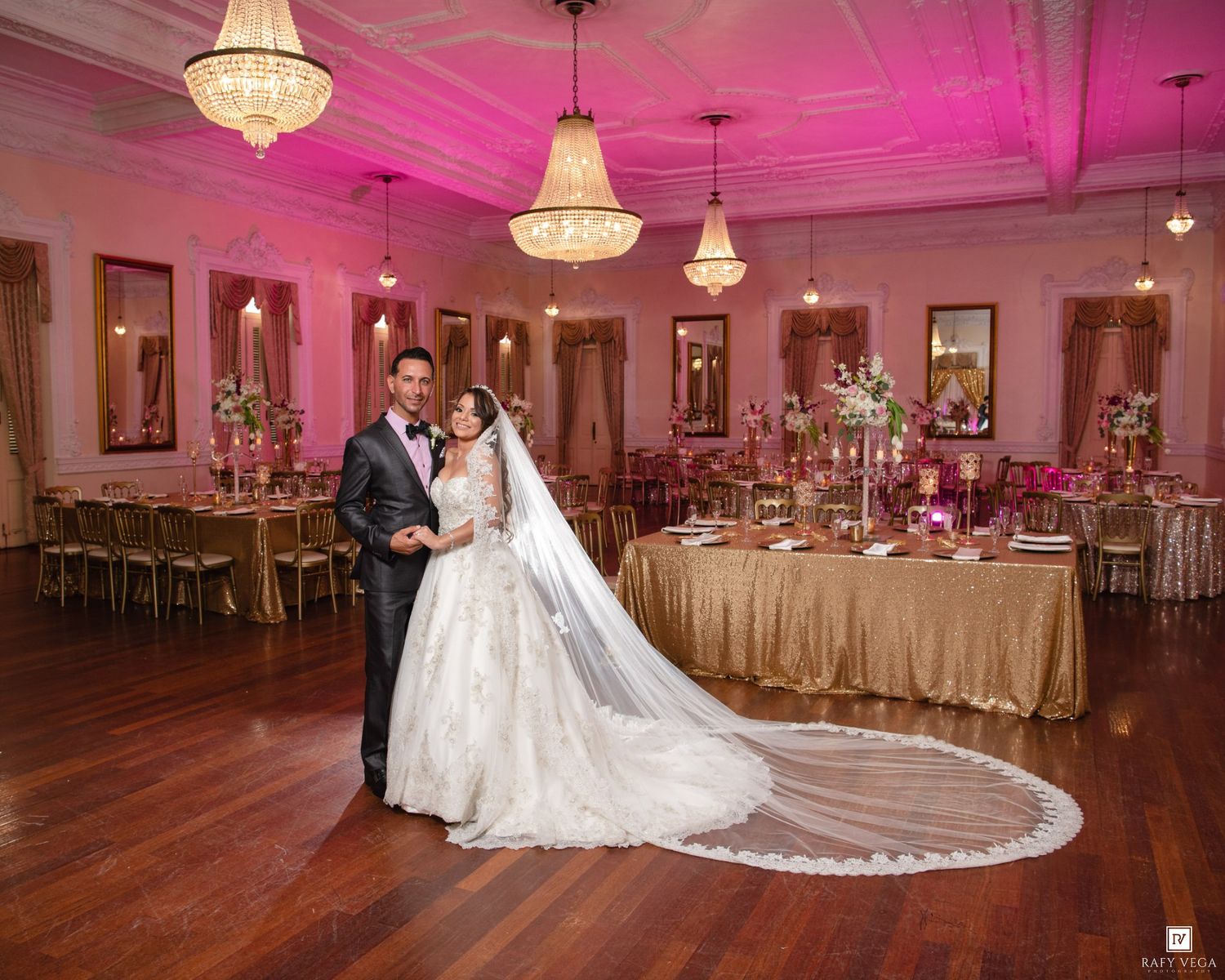 Bride and groom sharing first dance in elegant pink-lit ballroom with chandeliers and gold reception tables.