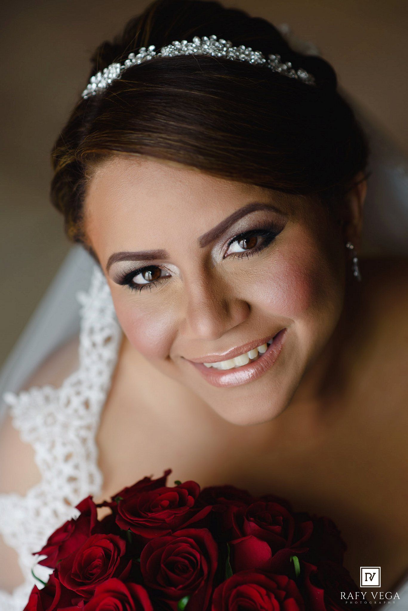 Smiling bride wearing a crystal tiara and lace dress holds red roses bouquet in a close-up wedding portrait.