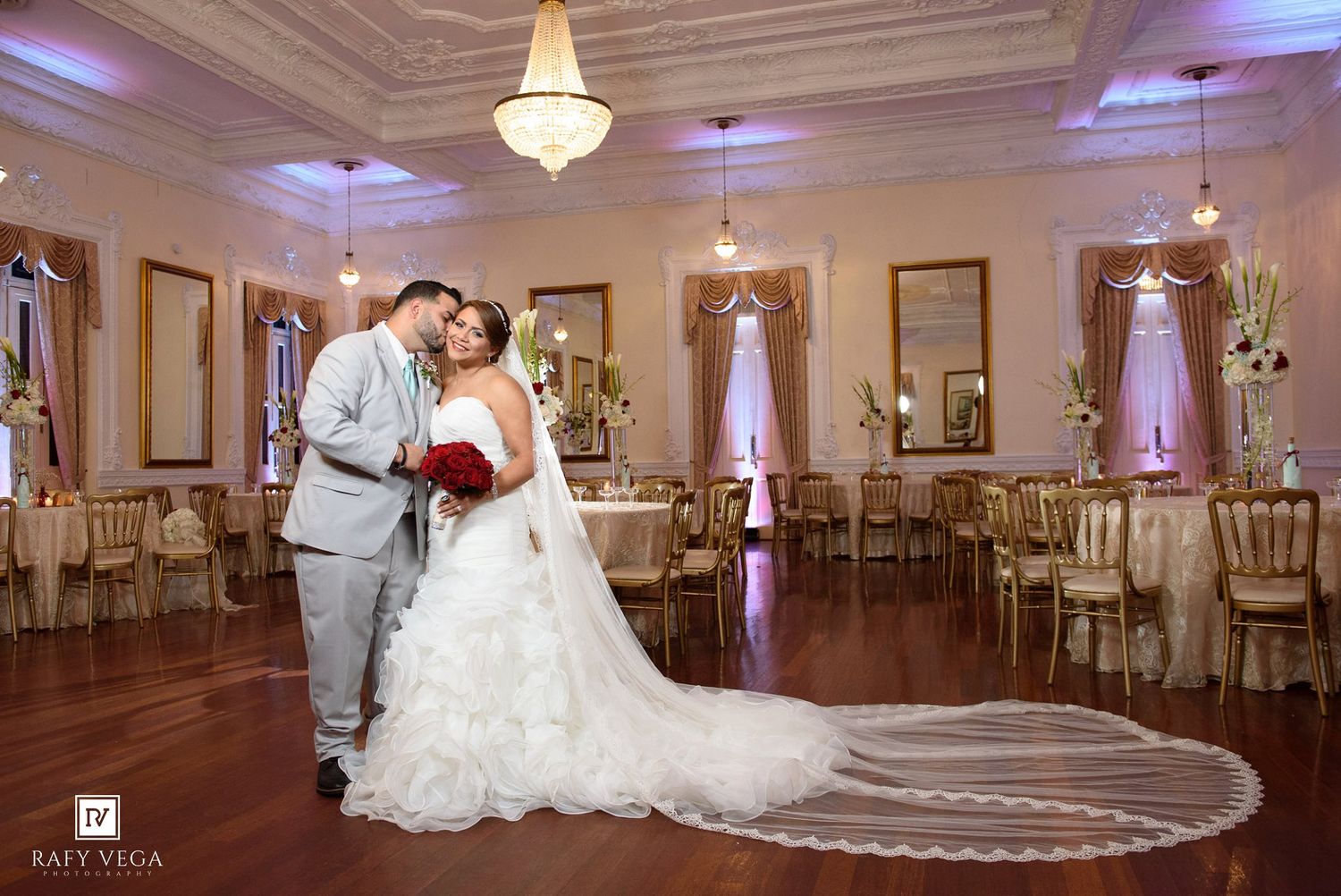 Bride and groom share romantic moment in elegant ballroom with gold chairs, mirrors, and cathedral-length veil.