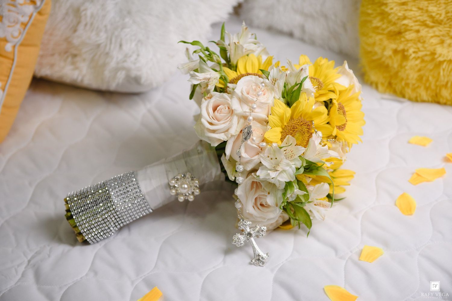 Yellow and white wedding bouquet with roses and rhinestone wrap on white quilted surface.