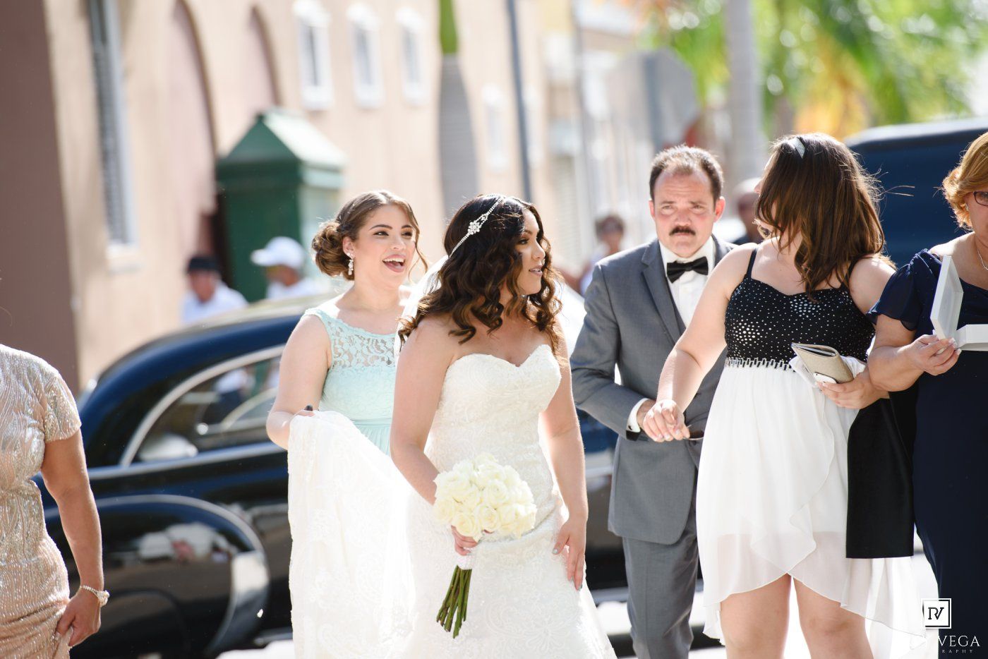 Bride in white gown holding bouquet walks with wedding party outside sunny venue.