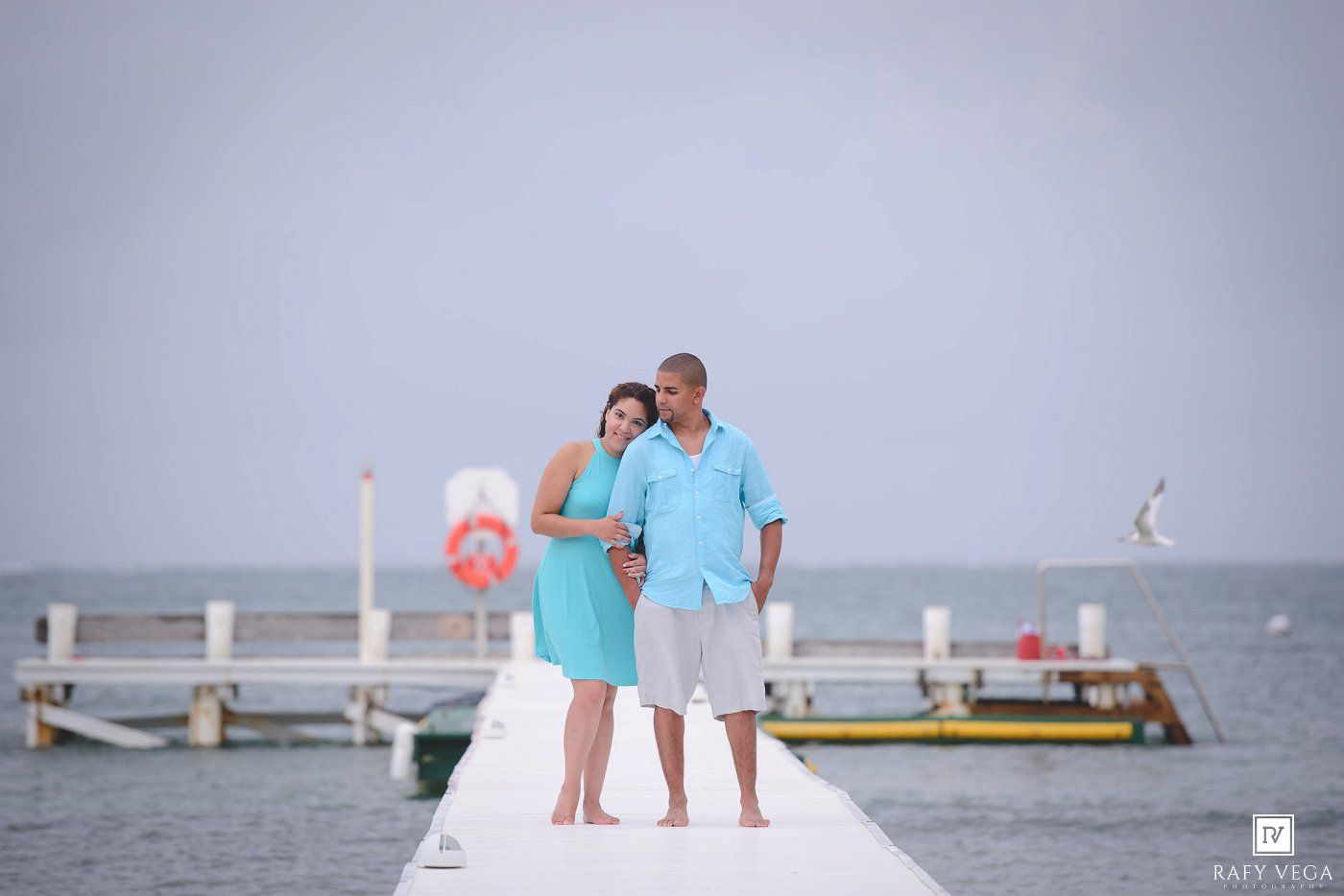 Couple posing on a white dock over calm turquoise ocean waters, woman in teal dress, man in light blue shirt.