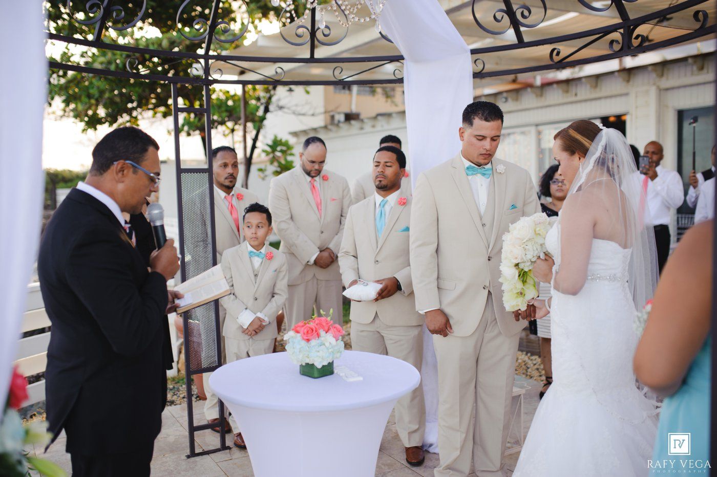 Groom in tan suit with bow tie stands at outdoor wedding altar with groomsmen and officiant.