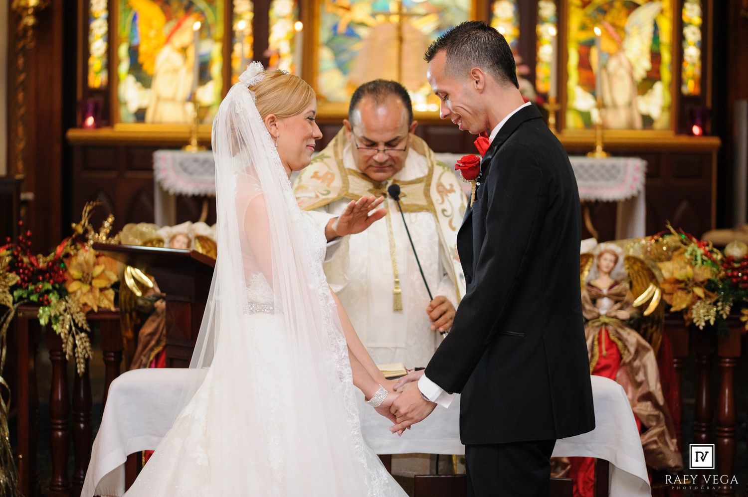 Bride and groom hold hands during wedding ceremony at ornate church altar with officiant in white robes.