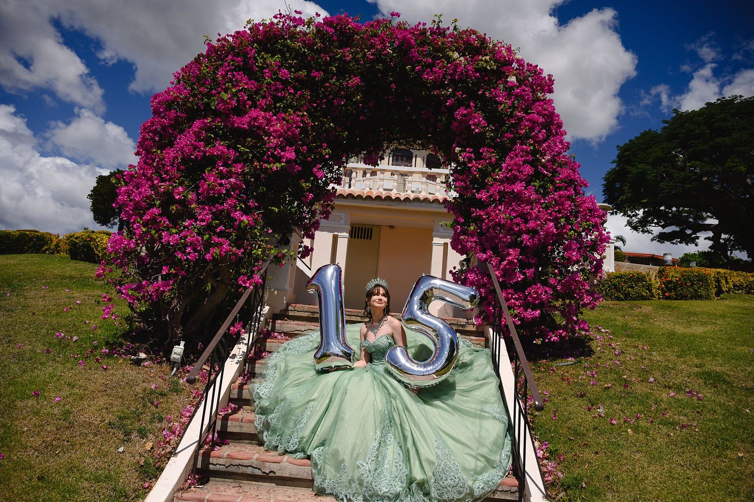 Girl in green quinceañera gown holds '15' balloons under floral arch with pink bougainvillea blooms.