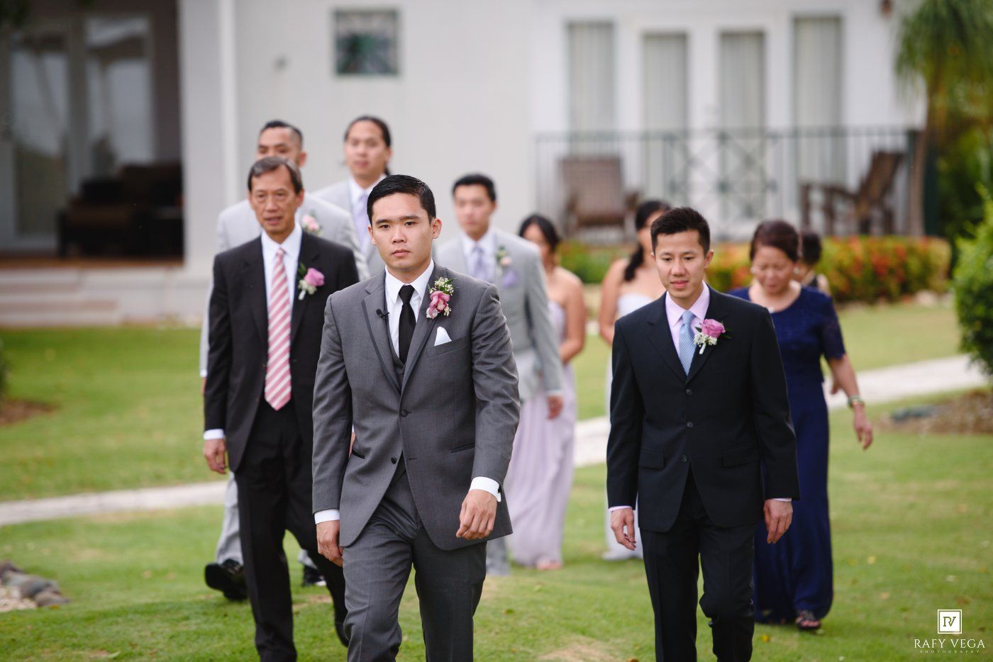 Groom and groomsmen in gray suits walk outdoors at a tropical wedding venue with guests following behind.