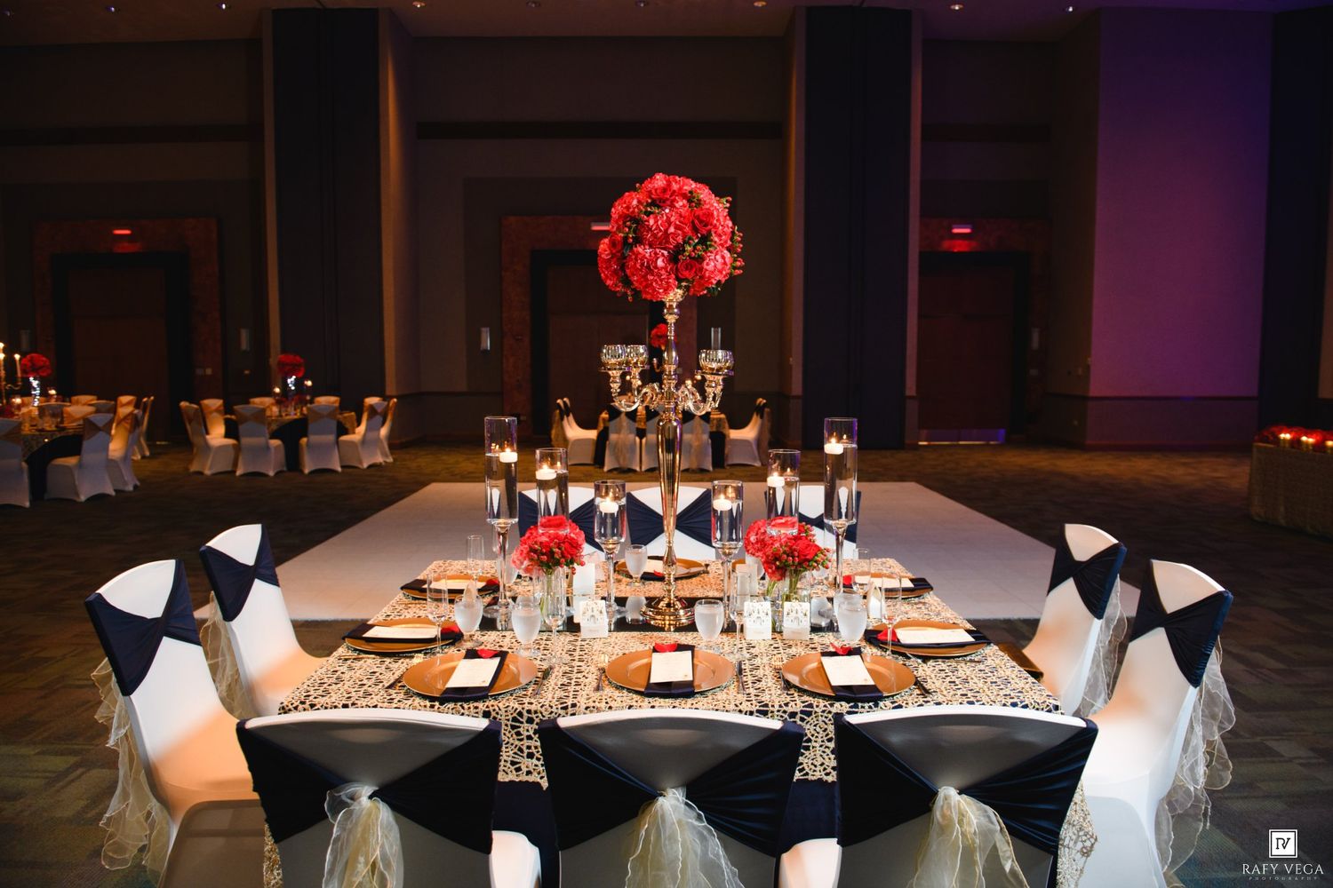 Elegant wedding reception with navy chair sashes, red floral centerpieces, candelabras, and gold accents on round tables in a ballroom.