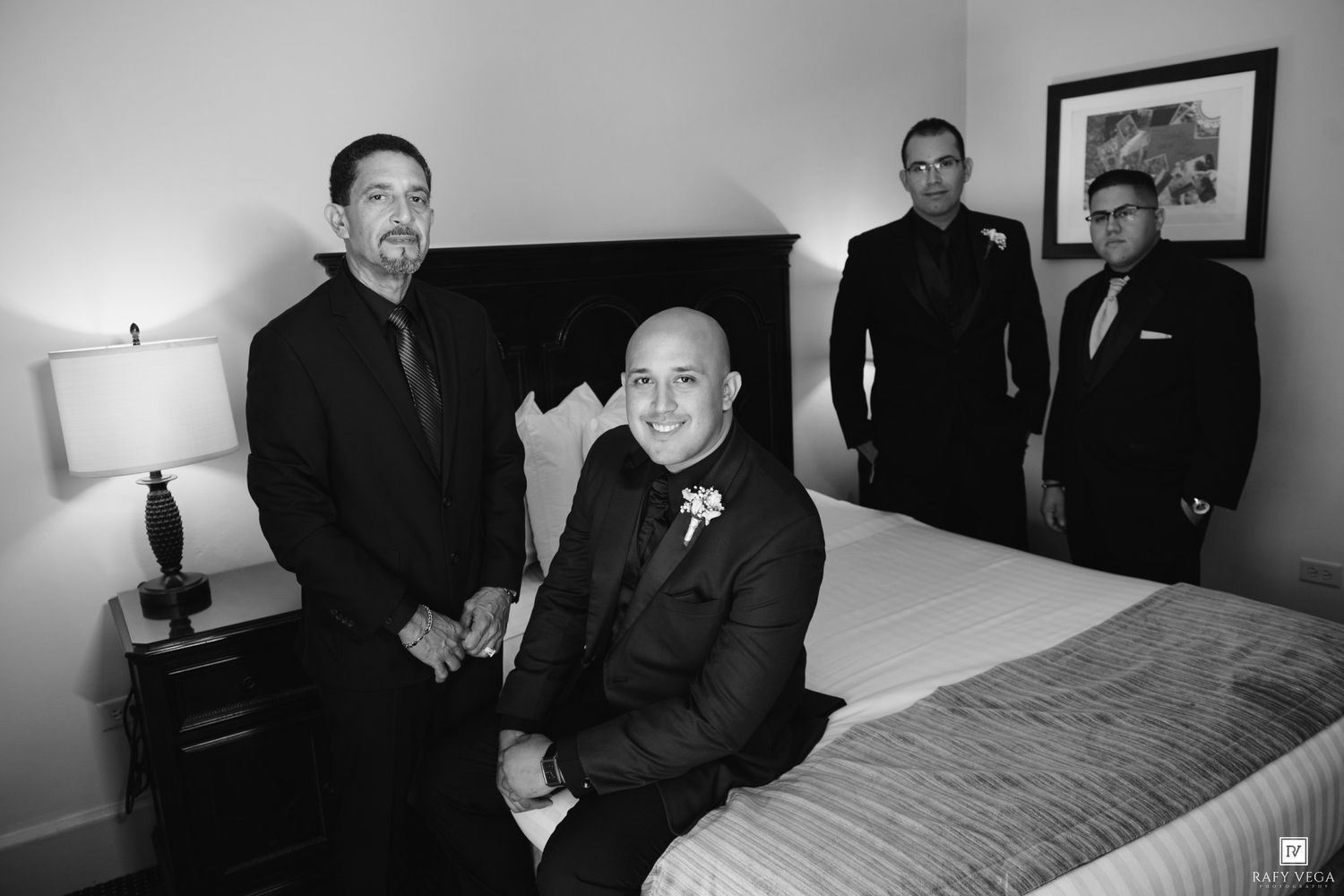 Black and white photo of a groom and three groomsmen in dark suits posing in a hotel room before a wedding.