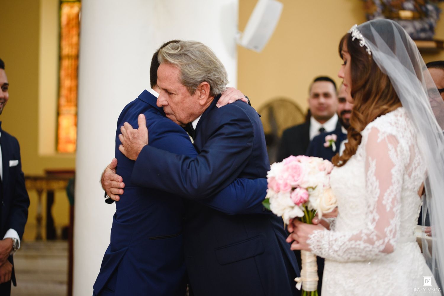 Two men in blue suits share an emotional hug at a wedding altar as a bride in lace gown holds pink bouquet.