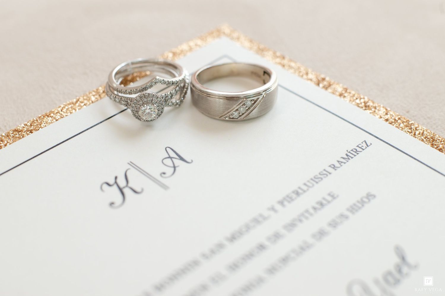 Two silver wedding rings resting on a elegant wedding invitation with gold glitter border and monogram initials.