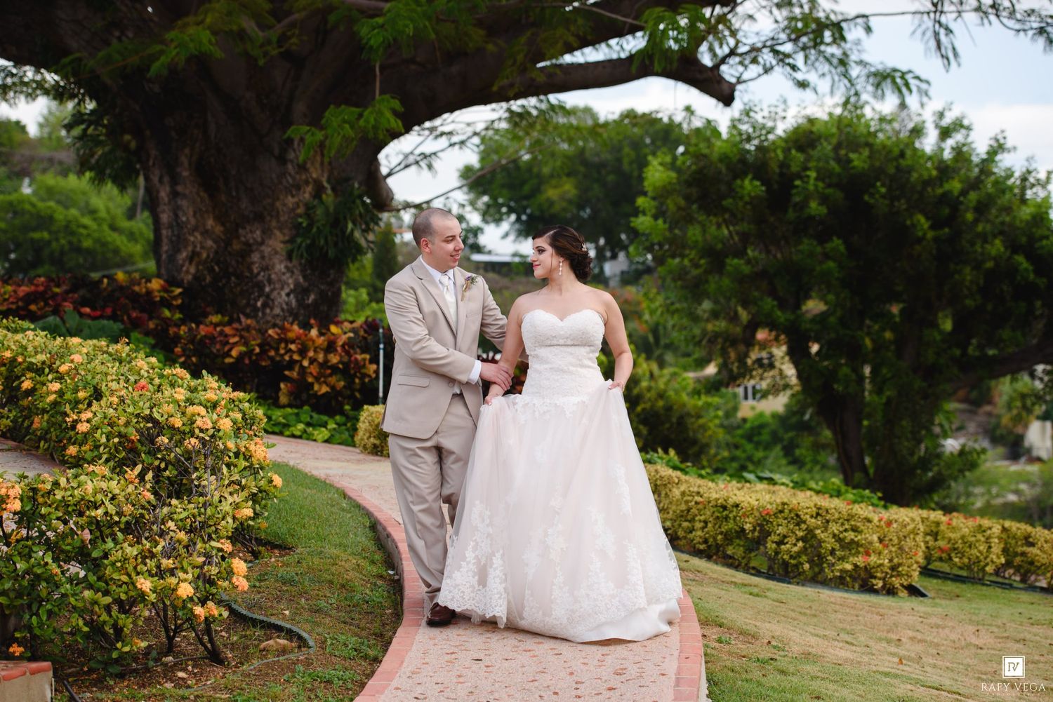 Bride in white gown walks with groom in tan suit through lush garden pathway on their wedding day.