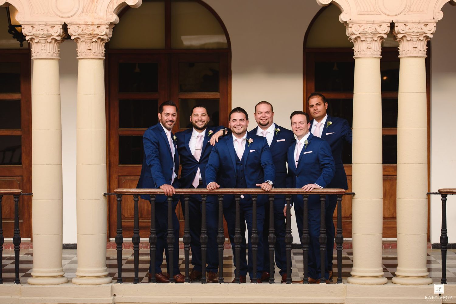 Six groomsmen in navy blue suits posing on a balcony with white columns at an elegant wedding venue.