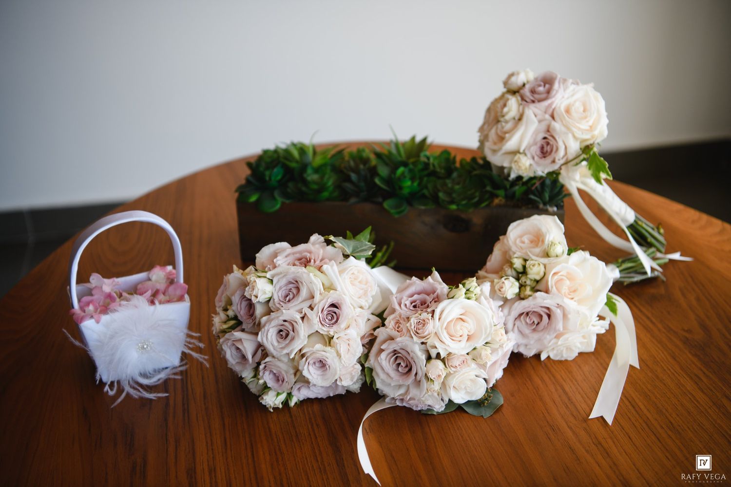 Elegant wedding bouquets of blush and mauve roses arranged on a wooden table with a floral wreath and flower girl basket.