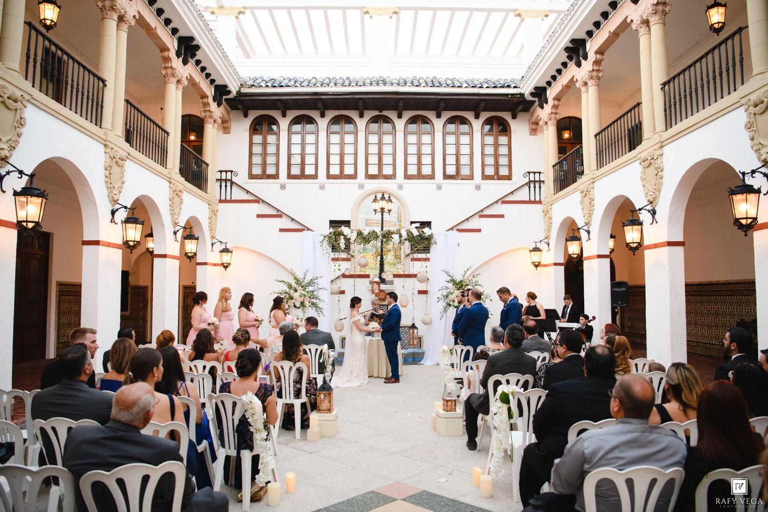 Elegant indoor wedding ceremony in a white Spanish-style courtyard with arched columns and seated guests.