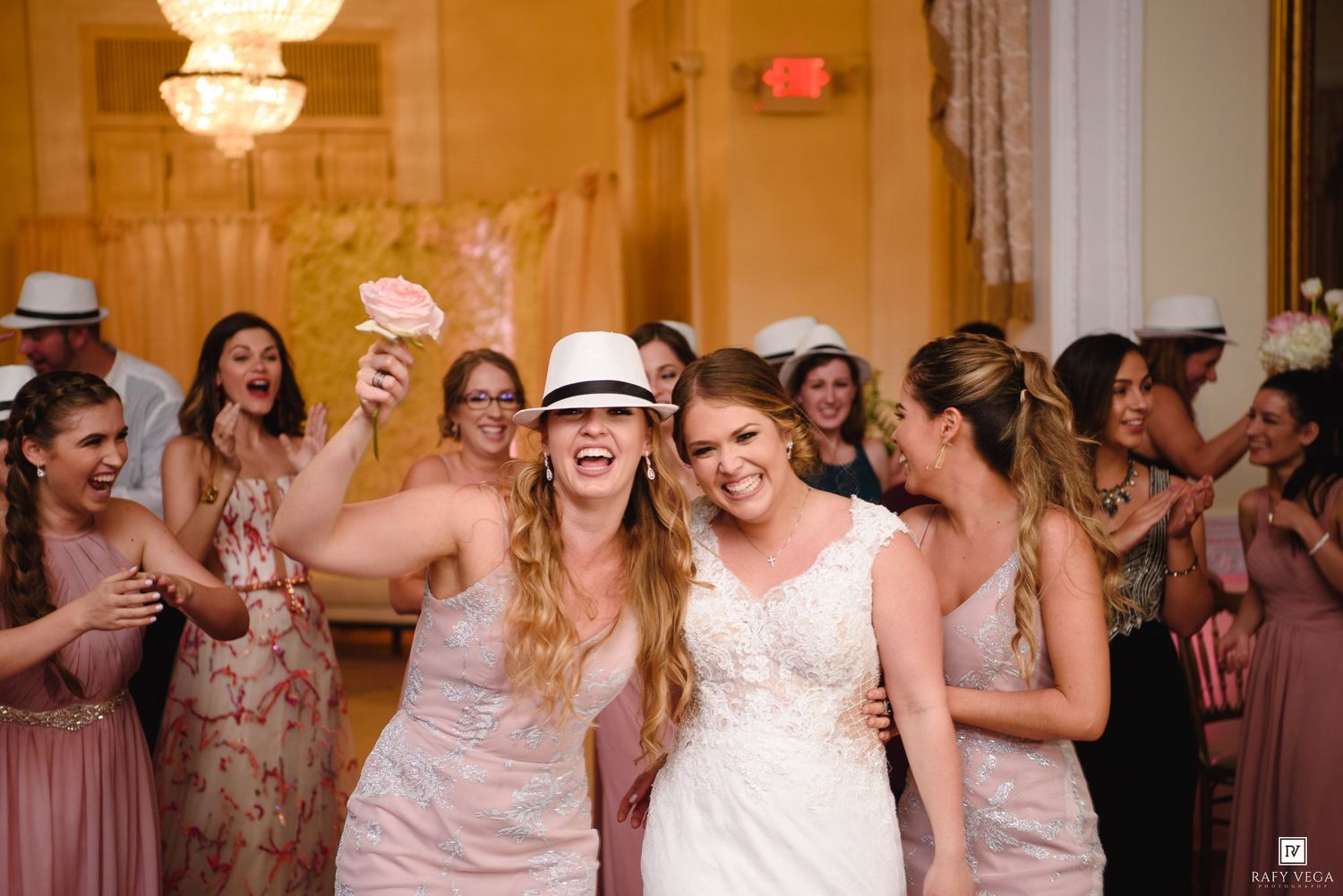 Bride and bridesmaid dancing joyfully at elegant wedding reception, surrounded by guests in pink dresses.