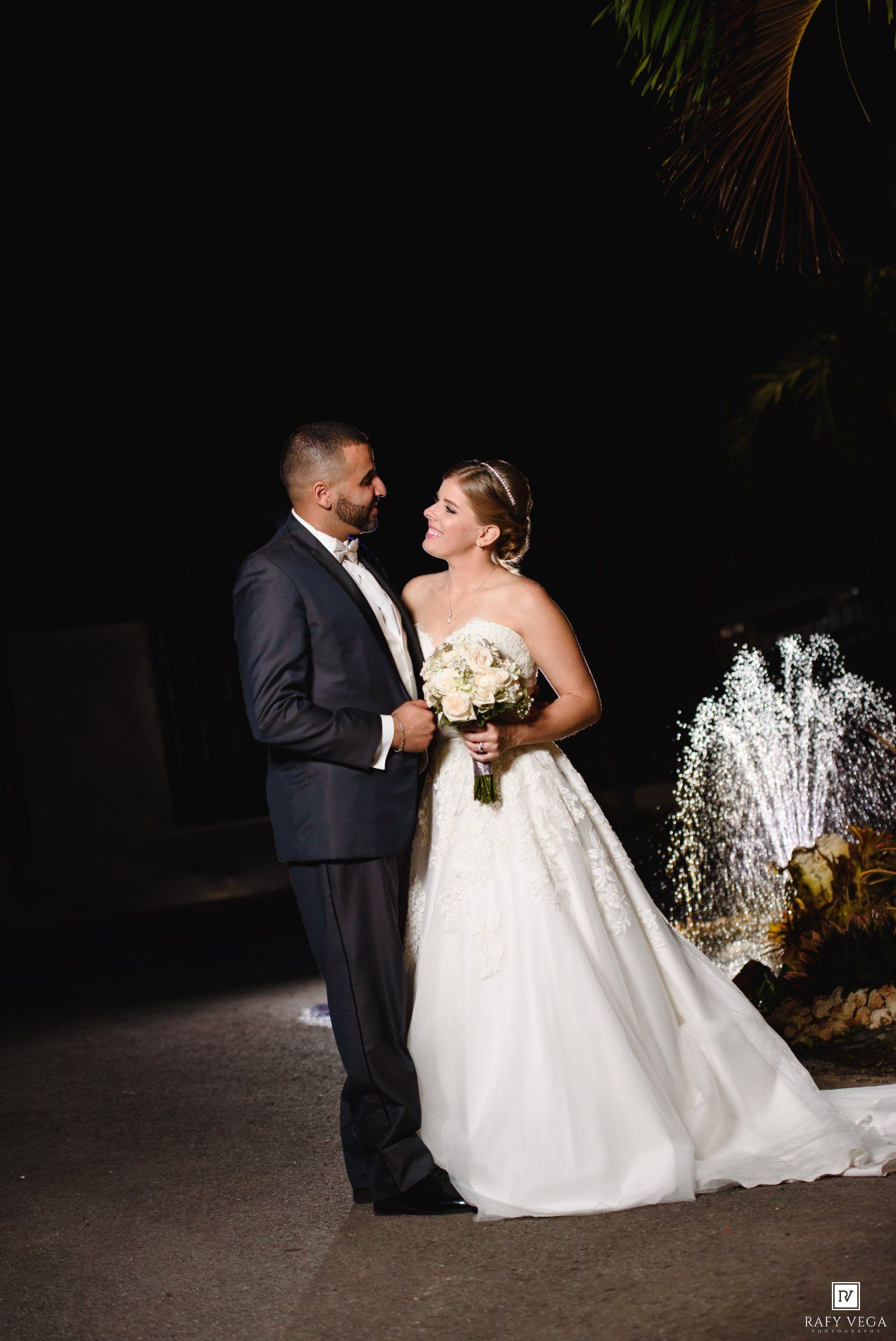 Bride and groom share a romantic moment outdoors at night, with a fountain and palm trees in the background.