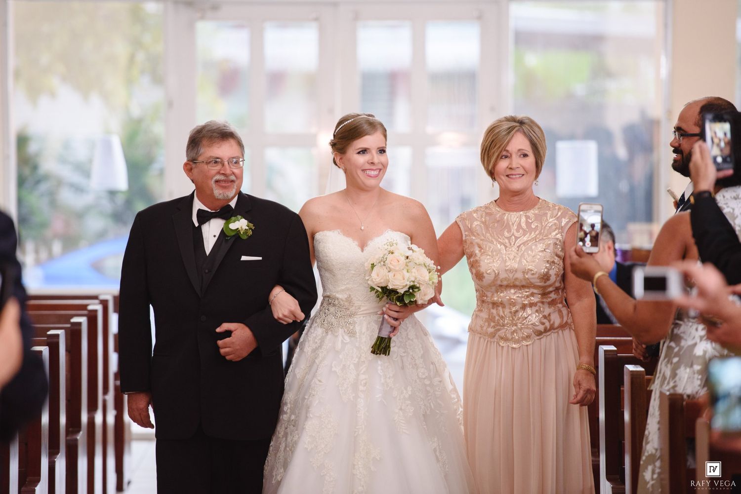 Bride walks down church aisle with parents on her wedding day, holding white bouquet in elegant ballgown.