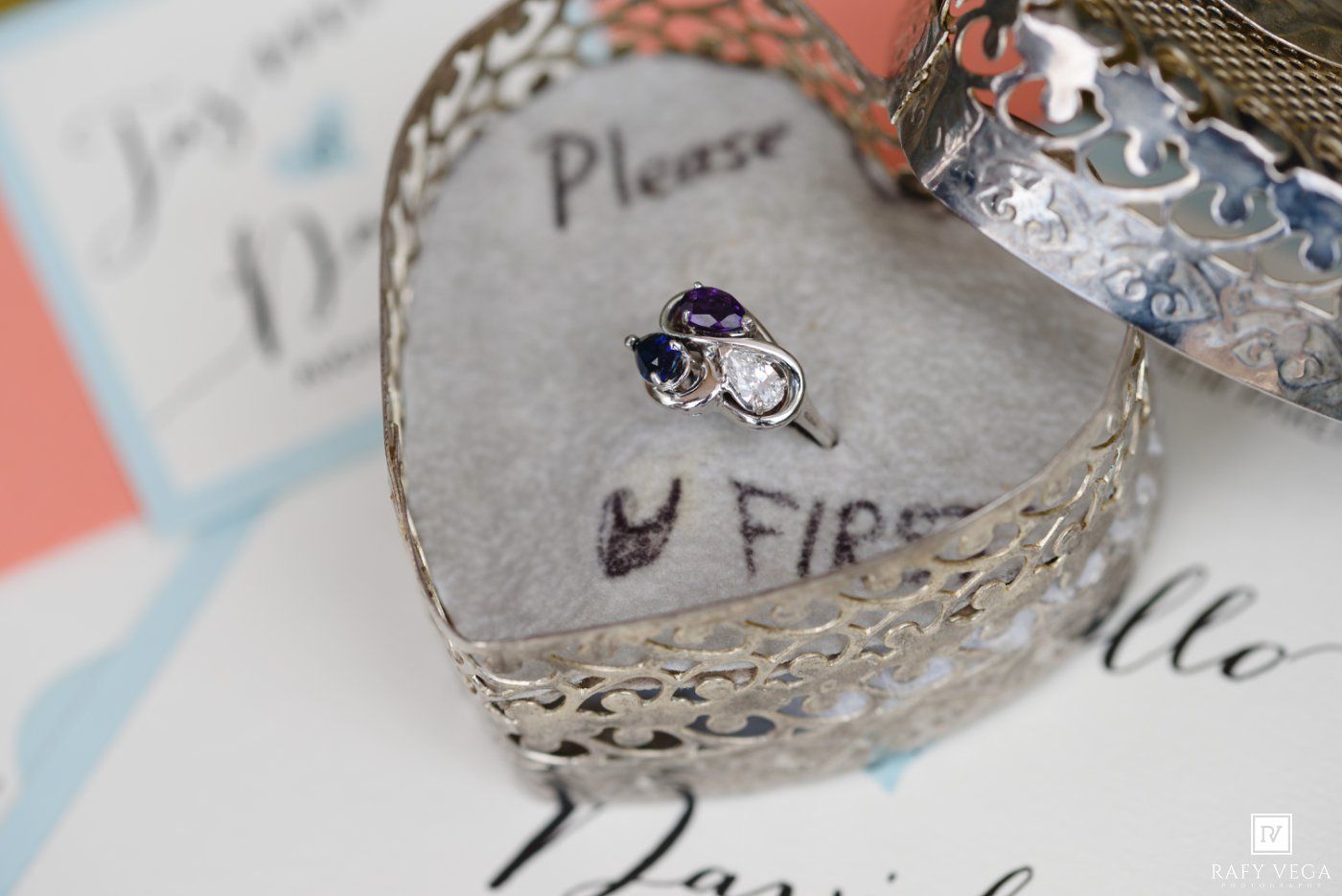 Silver filigree heart dish holding an amethyst engagement ring and wedding band atop elegant wedding invitation stationery.