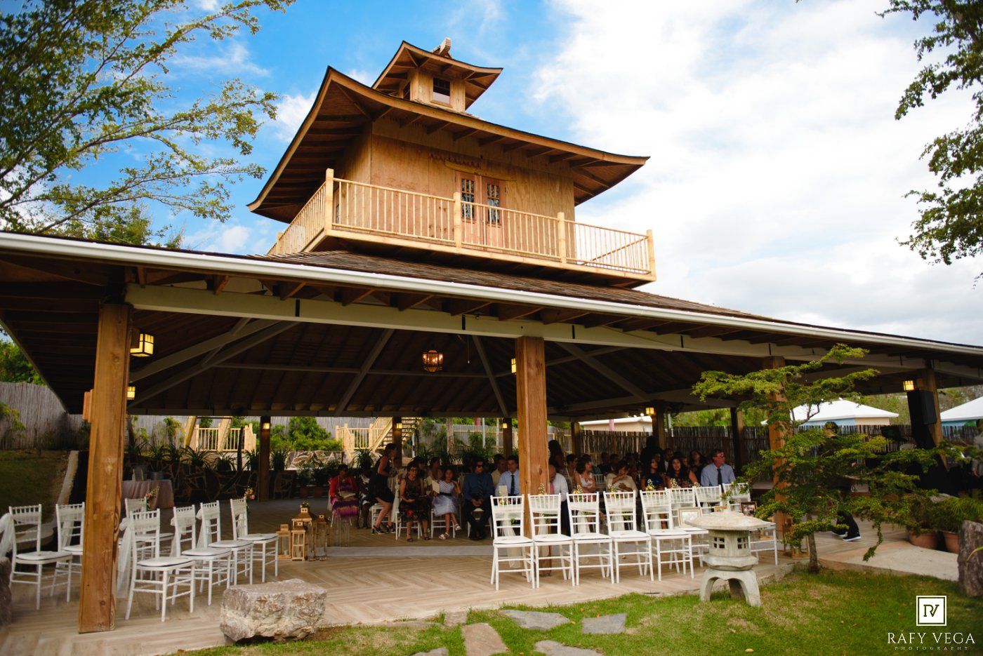 Elegant outdoor wedding venue with white chairs under a wooden pavilion featuring a Thai-style tower surrounded by lush greenery.
