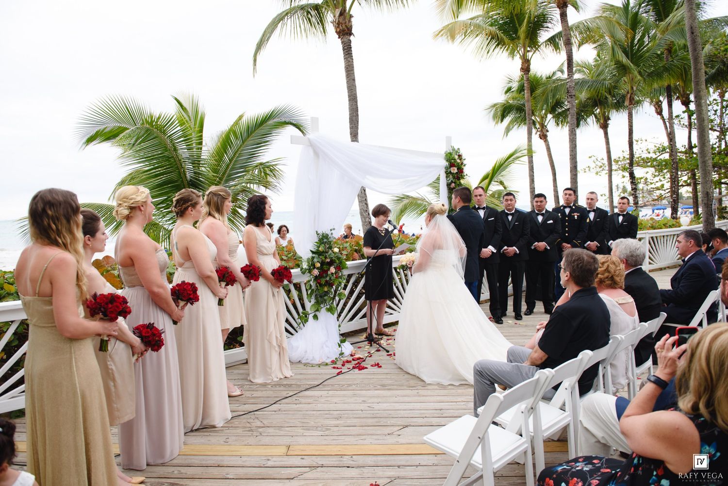 Outdoor beach wedding ceremony on a wooden deck with bridesmaids in blush gowns, groomsmen in black, and palm trees.