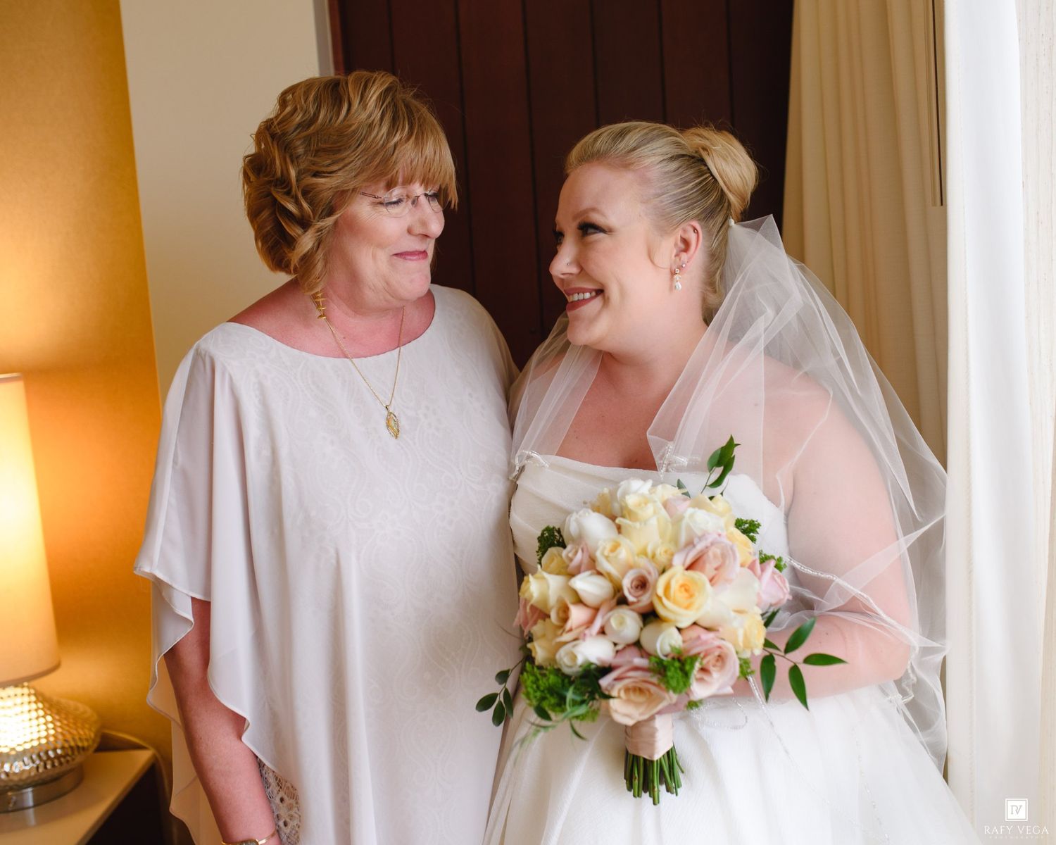 Bride in white gown holding pastel bouquet smiles with older woman in white dress near window.