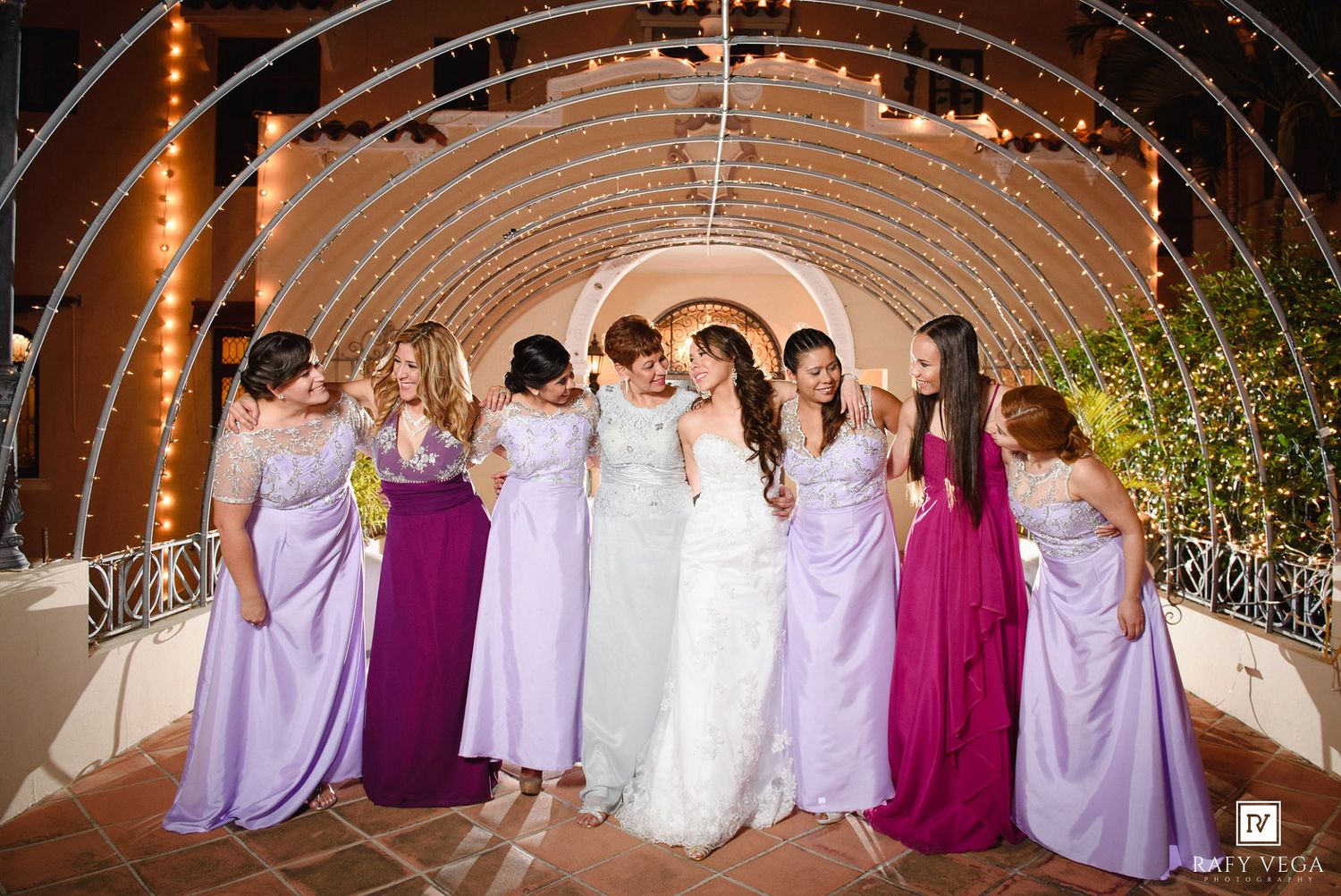 Bride and bridesmaids in purple gowns pose under a glowing arch at an elegant outdoor wedding venue.