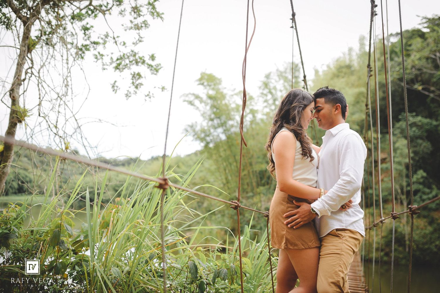 Couple sharing a romantic moment on a rustic swing bridge surrounded by lush greenery and a serene river.